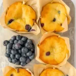 Blueberry Protein Muffins: 9g Protein, Irresistible Snack You’ll Love 6 Golden blueberry muffins in parchment liners, one broken open with fresh blueberries.