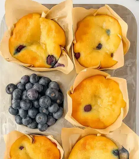Golden blueberry muffins in parchment liners, one broken open with fresh blueberries.