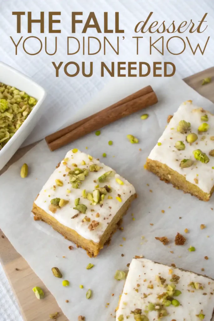 Pistachio Pumpkin Bars – The Fall Dessert You Didn’t Know You Needed 2 iced pumpkin pistachio cake squares on parchment with cinnamon sticks and crushed pistachios